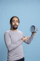 Punctual person with casual wear pointing at a wall clock on camera, looking at the hour and minutes on a watch against blue background. Young adult with long curly hair and beard is on time. photo