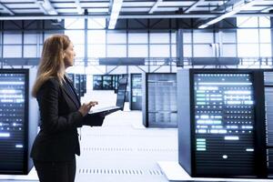 Computer scientist in data center checking rigs and hardware, doing checkup. Woman walking in high tech facility, planning and executing server hub migrations and upgrades using notebook photo