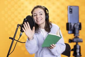 Happy media star waving hand while filming vlog at work, reading book to record audiobook. Cheerful woman doing content creation, recording novel, studio background, doing greeting gesture photo