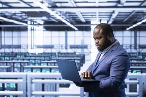 Worker in data center using laptop to configure network infrastructure such as routers, switches and firewalls. Data researcher working in server room, looking for technical malfunctions photo