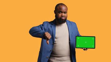 Upset african american man shows thumbs down sign gesturing holding isolated screen tablet, disapproving. BIPOC person does rejection hand gesture regarding mockup device, studio background, camera B photo