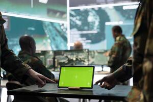 Two servicemen working in team with a green screen mockup, using a tablet to help troopers in power base command center. Soldiers examine real time information on device. Close up. photo