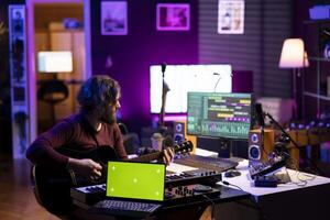 Sound engineer playing guitar next to laptop showing greenscreen display, recording songs with mixing console and daw software interface. Producer composes new music on acoustic instrument. photo