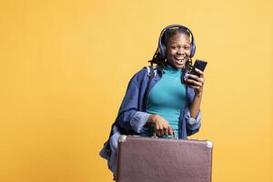 Portrait of overjoyed teenager reading messages on phone, receiving good news while travelling with suitcase, listening music. Woman excited by SMS on smartphone, using headphones, studio background photo