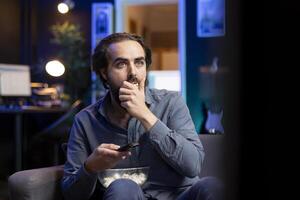 Cord cutter at home looking in disbelief at TV displaying thriller film, stuffing mouth with popcorn. Stupefied person at home watching suspenseful movie on VOD channel, perplexed by plot twist photo