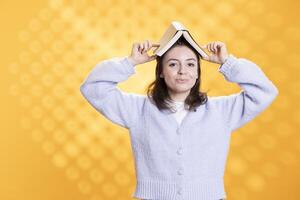 Portrait of happy woman placing book on head, enjoying reading hobby for relaxation purposes. Upbeat bookaholic person having fun wearing novel as hat, enjoying leisure time, studio background photo