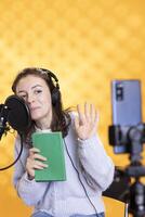 Influencer waving at phone camera, saluting followers, filming herself reading book, creating audiobook, studio background. Influencer greeting audience, doing vlog while recording audio novel photo
