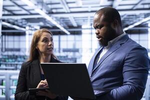 Employee in server hub showing manager recovery plan on laptop, monitoring energy consumption. Coworkers doing brainstorming in data center, making sure rigs are functioning optimally photo