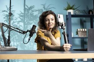 Cheerful woman using smartphone on selfie stick to capture footage, presenting professional microphone. Tech content creator showing studio mic to fans, filming with mobile phone photo