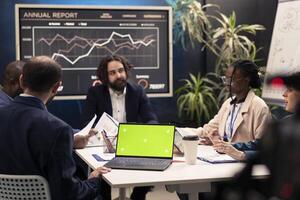 Startup team working with greenscreen on laptop at a conference table, examining promotional products to ensure sales growth. PR specialists collaborating on enterprise development. photo