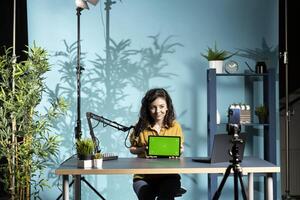 Jolly woman in studio filming chroma key tablet review for tech enthusiasts. Cheerful online star hosts technology internet show, unboxing green screen digital portable device photo