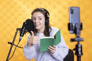Smiling influencer doing thumbs up at phone camera, filming reading book, creating audiobook, studio background. Influencer showing positive hand gesture, doing vlog while recording audio novel photo