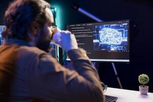 Programmer drinking coffee and doing maintenance on artificial intelligence neural networks computerized model. Tech support expert enjoying hot beverage while checking up on AI systems photo