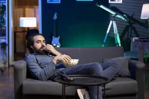 Relaxed person lounged on couch using TV to binge series on subscription based streaming services, enjoying bowl of popcorn. Man laying down watching VOD shows on television display, eating snack photo