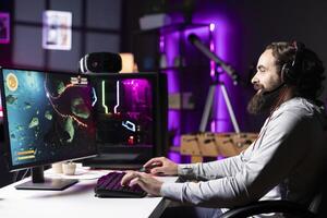 Smiling man playing videogames live while chatting with online audience from RGB lit living room. Cheerful pro gamer streaming game while talking with subscribers using headphones mic photo