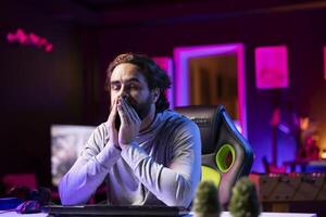 Man feeling tired after working all day, struggling to stay awake while playing games on computer in rgb lit home. Player trying to relax by competing in esports competitions but falling asleep photo