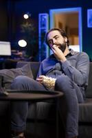 Fatigued man watching shows on TV and munching on popcorn, paying close attention to screen. Tired sedentary man sitting in pajamas all day devouring snacks and consuming mindless entertainment photo