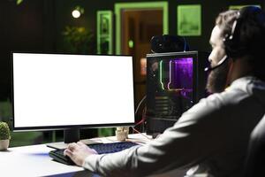 Man wearing headphones playing games on isolated screen gaming PC at computer desk, enjoying day off from work. Gamer battling enemies in online multiplayer shooter on mockup monitor photo