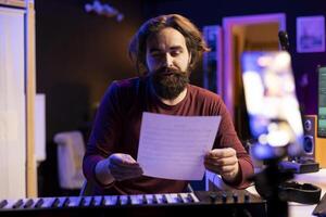 Skilled songwriter explaining music sheet with notes on online lesson vlog, doing internet classes in his home studio. Young artist teaching people how to sing, midi controller keys. photo
