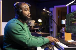 African american artist composer playing electronic keyboard to record acoustics on professional software and add sound effects. Audio engineer editing soundtracks with stereo console and tools. photo