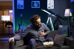 Sedentary man at home feeling terrible pain in chest while watching TV on couch after lack of activity. Person getting painful cardiac stroke from consuming unhealthy snacks all day, gripping chest photo