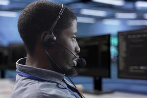 Employee working in call center sitting at PC desk and providing outsourcing services for data centers. Man at work talking in headphones microphone with on field admins in server farm, close up shot photo