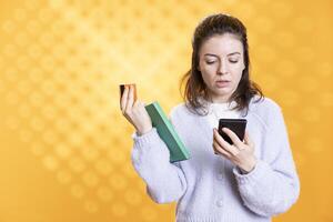 Woman adding credit card information on smartphone while holding book, studio background. Person with academic textbook in hand purchasing more educational material using phone photo