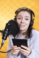 Portrait of smiling woman reading ebook from ereader, recording audiobook using microphone, studio background. Jolly narrator using mic to produce digital recording of novel, reading text from tablet photo