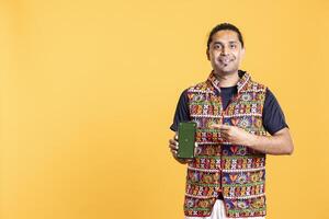 Portrait of influencer smiling while presenting green screen mobile phone, studio background. Jolly indian person holding copy space chroma key smartphone used for advertising brands, grinning photo