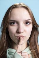 Stern teenager doing shushing hand gesturing, irritated by loud noises. Extreme close up shot of girl placing finger on lips, doing quiet sign gesture, blue studio background photo