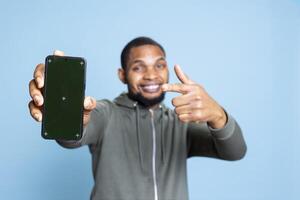 Portrait of african american person pointing at isolated copy space screen on smartphone, standing against blue background. Young man in casual clothes holding mockup phone device in studio. photo