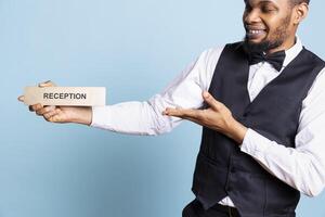Young bellhop pointing at an indicator for the reception area front desk, working at a five star hotel and guiding clients to all the resort amenities. Bellboy doorkeeper with suit and tie. photo
