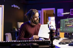 Young singer pointing at music sheet to explain notes on synthesizer, teaching people how to play piano electronic keyboard in home studio. Artist performer playing midi controller keys, online vlog. photo