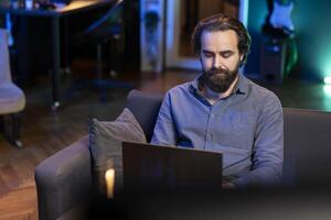 Man working on laptop while watching live sports event on TV in living room. Teleworking employee at home typing on notebook, solving business tasks and keeping football match in background photo