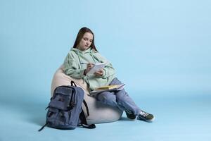 Dutiful young girl sitting on beanbag, reviewing school notes to gain knowledge, studio background. Learner with notepad in arms reading college courses, doing research for exams photo