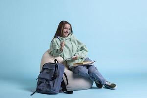 Portrait of jolly girl laughing while sitting on beanbag and writing on notebook, studio background. Overjoyed schoolgirl with notepad in arms feeling amused while preparing homework photo