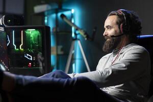 Happy gamer plays online multiplayer videogame with friends, speaking with them using headphones mic. Cheerful player talking with teammates, putting feet up, enjoying leisure time photo
