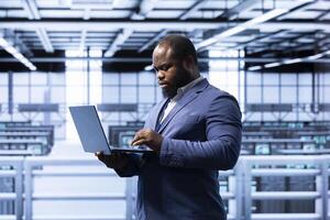 IT staff member in server hub using laptop to monitor energy consumption across supercomputer rigs. Computer scientist in data center making sure rackmounts are functioning optimally photo