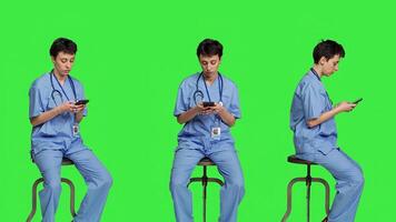 Nurse browsing online websites on smartphone app in studio, sitting on a chair against greenscreen backdrop. Medical specialist in scrubs using mobile phone and social media texting. Camera A. photo