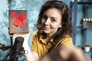 Joyful content creator holding present box, excited to do giveaway for subscribers, filming with selfie camera. POV shot of media star happy to offer packed gift with ribbon to viewers photo