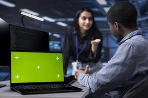 Focus on isolated screen laptop used by team in blurry background reconfiguring server hub to optimize it for data processing. Close up of chroma key notebook used by workers overseeing data center photo