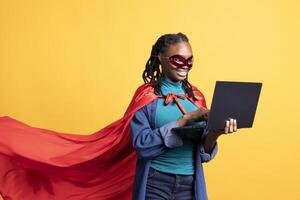 Smiling african american girl portraying superhero hiding identity, working on laptop, studio background. Jolly woman posing as hero in costume, solving tasks on notebook, covering face with mask photo