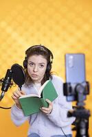 Influencer wearing headphones filming herself reading book with cellphone on tripod, creating audiobook, studio background. Woman doing vlog with producing recording on novel using professional mic photo
