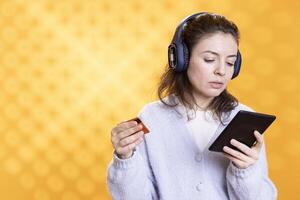 Woman reading ebook on ereader and listening music purchasing books online. Person with tablet in hands looking to buy digital novel with credit card, hearing songs in headphones, studio background photo