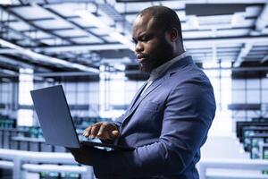 Engineer in server room establishment installing newly ordered storage units using laptop. African american man working in data center facility housing storage rackmounts photo