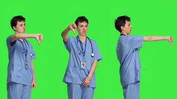 Displeased medical assistant shows thumbs down in studio, presenting her disapproval and disagreement with dislike symbol against greenscreen. Nurse feeling unhappy with rejection sign. Camera B. photo