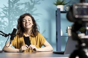 Smiling tech expert using professional camera to film technology review of new cellphone, doing unboxing . Happy content creator showing phone box to subscribers, recording with high tech gear photo