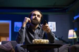 Man using remote control to switch channels on television screen while eating popcorn. Person scrolling through streaming content, looking for interesting movie, enjoying snacks photo