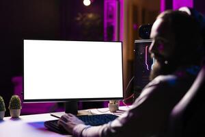 Man in dimly lit apartment playing games on isolated screen gaming PC at computer desk, enjoying day off from work. Gamer battling enemies in online multiplayer shooter on mockup display photo