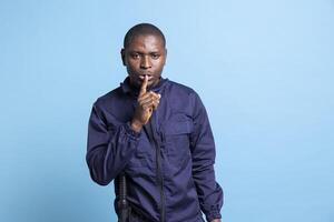 Man dressed in security agent uniform presents a mute gesture in studio, asking for silence and a quiet moment with a finger over lips. Young adult guard showing a hush sign, blue background. photo